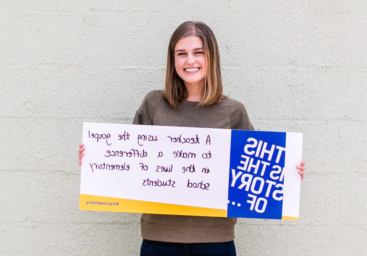 A female undergraduate displaying a sign that states, "This is the story of a teacher using the gospel to make a difference in the lives of elementary school students."